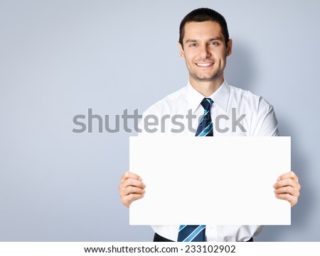 Portrait of happy smiling young businessman showing blank signboard, with copyspace area for text or slogan, against grey background