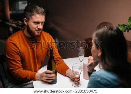 High-angle view of young man pouring wine into glass during talking with wife sitting together at festive table. Love couple celebrating anniversary or Valentine day having romantic dinner
