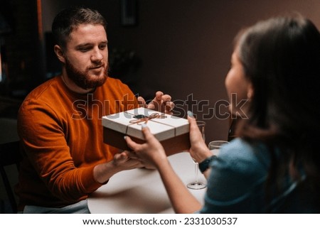 Happy young man receiving present from loving wife enjoying romantic dinner date sitting at table with candles on birthday or Valentines Day. Pretty young woman giving wrapped box with gift to husband