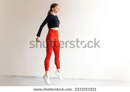 Active healthy woman doing cardio exercises and jumping in front of windows of white studio. Young energetic female dressed in tight sports clothes training legs muscles. Royalty-Free Stock Photo #2331051031