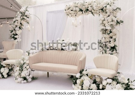 Beautiful decor for the wedding ceremony in the form of a sofa and white flowers on the ceremonial arch. Light furniture in the form of a sofa and chairs in a white tent is decorated with white roses Royalty-Free Stock Photo #2331072213