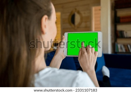 Back view of young female holding horizontal tablet with green screen sitting in room. 