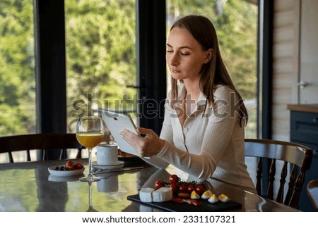 Woman uses a digital tablet or browses the Internet, sits at a table with fruit and cheese and a cup of coffee against the background of the kitchen interior, dressed in home clothes. 