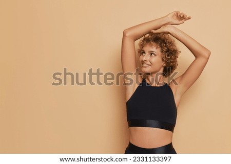 Slim girl with short curly hair stands aside on peachy background, looks at empty space with smile, holds hands up on her head, lifestyle concept, copy space