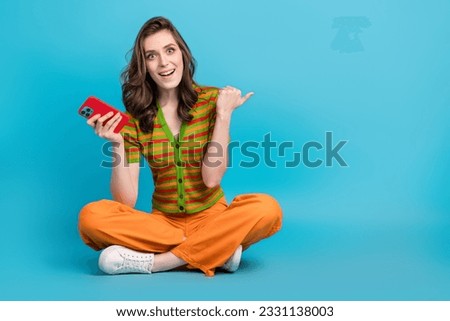 Full length photo of impressed girl wear striped shirt trousers hold smartphone directing empty space isolated on blue color background