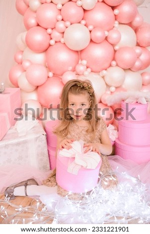 A beautiful little girl in a festive dress sits in pink and white balloons, with a box of gifts in her hands, and a luminous garland