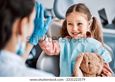 A candid emotional photo of a child sitting in a dental chair, holding a toy rabbit and cheerfully giving a high-five to the nurse. Children's dentistry.