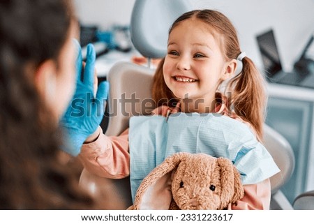 At the doctor's appointment. A candid emotional photo of a child sitting in a dental chair, holding a toy rabbit and cheerfully giving a high-five to the nurse. Royalty-Free Stock Photo #2331223617
