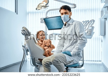Children's dentistry.Portrait of a dentist doctor in a medical gown and mask sitting and in the background a little female patient sitting in a chair and holding a toy.  Royalty-Free Stock Photo #2331223625