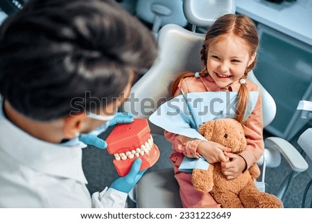 A little girl sits in a dental chair, holds a toy rabbit in her hands, laughs and looks at the dentist who shows her a model of the jaw with teeth. Children's dentistry. Royalty-Free Stock Photo #2331223649