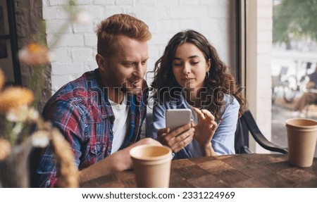 Positive young couple in casual clothes sitting at wooden table with cups of coffee and browsing mobile phone during break Royalty-Free Stock Photo #2331224969