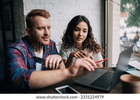 Millennial male and female bloggers analyzing web page during business cooperation in coffee shop, skilled graphic designers creating website together during collaborative distance job in coworking