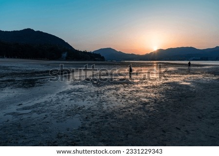 anoramic view over shoreline of Miyajima (Itsukushima) Island, Japan during low tide and dusk with two people on mudflat making pictures of the sun setting behind the mountains on other side of water