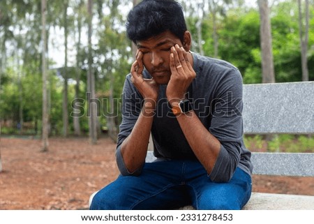 A depressed Indian man sits on a bench in a park, tears streaming down his face. He looks lost and alone, his head in his hands. Royalty-Free Stock Photo #2331278453