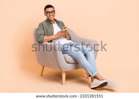 Full size photo of handsome positive man wear stylish shirt denim pants sit in armchair hold smartphone isolated on beige color background