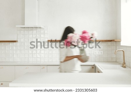 Blurred woman arranging peonies in vase on background of minimal white kitchen in new modern home. Motion image of housewife decorating house with flowers, housekeeping