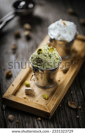 Pistachio and vanilla ice cream scoops in metal cups. Served on wooden tray with whole nuts. Italian gelato on rustic table.