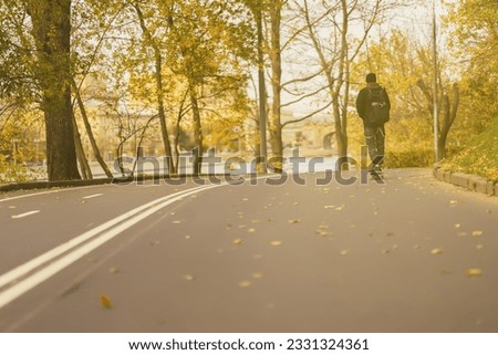 Sunny autumn day in park. Unrecognizable guy, skater back to us on skateboard, longboard skates along the paths among the many crumbling leaves. Concept youth leisure and sports, autumn