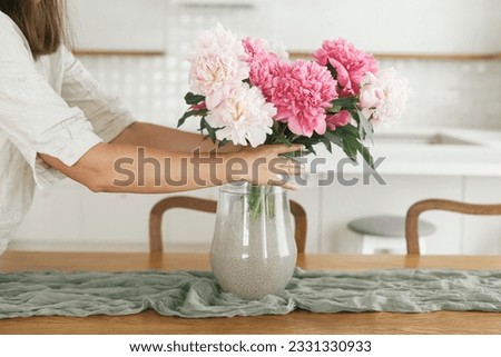 Woman arranging beautiful peonies in vase in new modern home. Young female decorating house with flowers on background of minimal white kitchen, hands with flowers close up