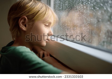 Portrait of a sad preteen boy standing at the window. Child watching the rain outside. Post-traumatic disorder. Accommodation of grief. Experiencing loss. Fear. Depression. Stress. Upset offended kid. Royalty-Free Stock Photo #2331333103