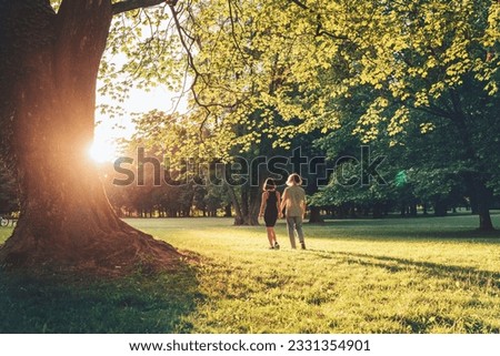 Young long-haired teenage boy with girlfriend walking hand in hand under huge tree during an outdoor walk in a summer city park at sunset time. First love, modern teenagers relations concept image.