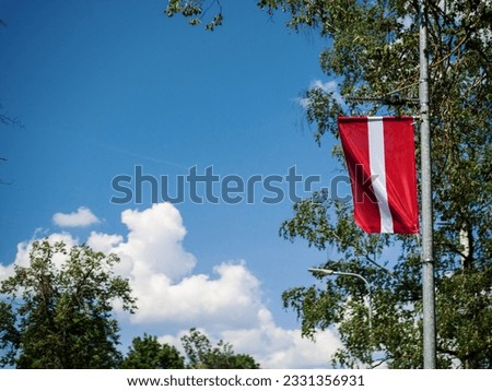 Latvian National flag on a metal pole. Town decoration for special military or government event. Blue cloudy sky in the background.