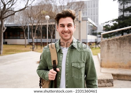 Portrait of a young male student standing up and looking at camera. Guy smiling and hold a backpack outdoors the college. Cheerful Gen z Caucasian boy in a university campus