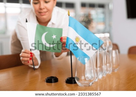 Little flag of Argentina on table with bottles of water and flag of Pakistan put next to it by positive young woman in meeting room