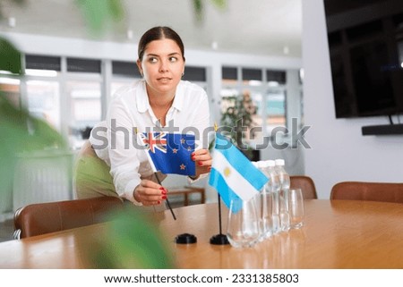 Positive young woman putting little flag of Australia on table next to the flag of Argentina and bottles of water in conference room