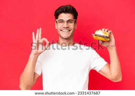 Young Caucasian man holding a burger isolated on red background showing ok sign with fingers