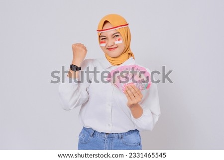 Smiling young Asian Muslim woman wearing hijab Celebrate Indonesian independence day on 17 August while holding money Indonesian rupiah banknotes isolated over white background Royalty-Free Stock Photo #2331465545