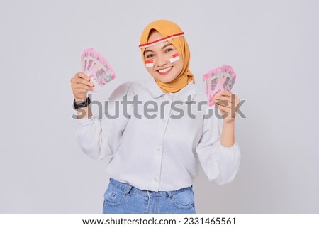 Smiling young Asian Muslim woman wearing hijab Celebrate Indonesian independence day on 17 August while holding money Indonesian rupiah banknotes isolated over white background Royalty-Free Stock Photo #2331465561