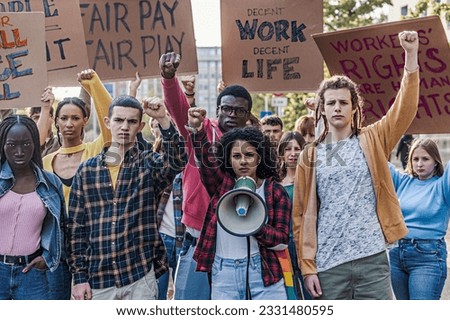 Multiethnic Youth Protesting for Decent Work and Life - A group of multiethnic youth protesting for fair pay and work access, raising fists. A curly-haired woman shouts slogans through a megaphone Royalty-Free Stock Photo #2331480595