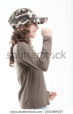 Smiling girl in military green, camouflage cap, and jeans, dances joyfully. Her long curls sway with her ballet-like movements on white backdrop