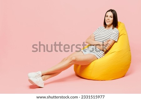 Full body young caucasian woman wears casual clothes t-shirt sit in bag chair hold hands crossed folded look camera isolated on plain pastel light pink background studio portrait. Lifestyle concept
