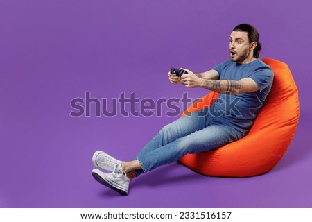 Full body young smiling man 20s wear basic blue t-shirt sit in bag chair hold in hand play pc game with joystick console isolated on plain purple background studio portrait. People lifestyle concept