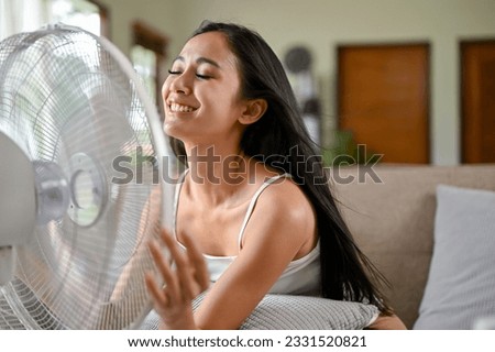 A happy and relaxed Asian woman is refreshing and cooling herself down with an electric fan while resting on a couch in her living room on a hot summer day. Royalty-Free Stock Photo #2331520821
