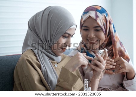 Pretty young Muslim woman with her older sister wears hijab sharing something on cell phone while sitting on sofa at home, talking and smiling 