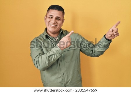 Hispanic young man standing over yellow background smiling and looking at the camera pointing with two hands and fingers to the side.  Royalty-Free Stock Photo #2331547659