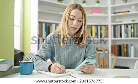 Young blonde woman student using smartphone writing notes at library university