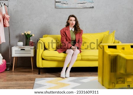 concept photography, beautiful well dressed young woman with wavy hair sitting on yellow couch, stylish house interior, vase with tulips, watching tv, posing like a doll, housewife lifestyle Royalty-Free Stock Photo #2331609311