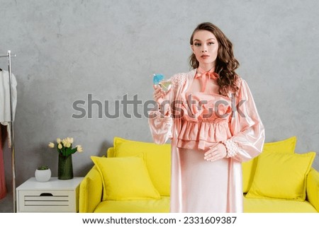concept photography, woman acting like a doll, domestic life, housewife in pink outfit with silk robe holding cocktail in glass, gesturing and standing near yellow coach in modern living room