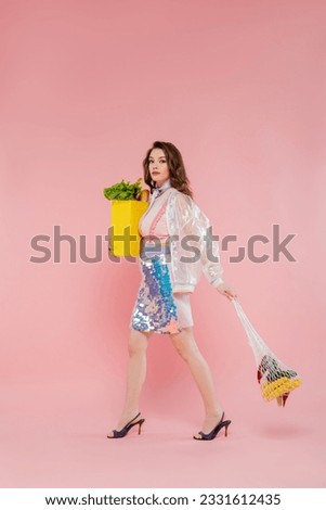housewife concept, happy young woman holding reusable string bag with groceries, stylish wife doing daily house duties, walking on pink background, looking at camera, fashionable