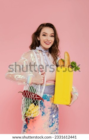 housewife concept, happy young woman holding reusable mesh bag with groceries, stylish wife doing daily house duties, standing on pink background, looking at camera, role play
