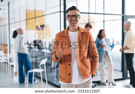 Positive adult male employee in casual wear and eyeglasses standing near colleagues while looking at camera with smile and showing thumb up Royalty-Free Stock Photo #2331613207