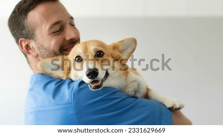 Friendly man vet in blue uniform cuddling, embracing pembroke welsh corgi dog, doctor carrying and playing with little dog after treatment, panorama, empty space Royalty-Free Stock Photo #2331629669