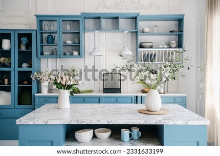 Marble countertop with vases and flowers in provence style apartment. Kitchen island and dining table with tableware. Blue furniture and white walls in classic interior design room. Royalty-Free Stock Photo #2331633199