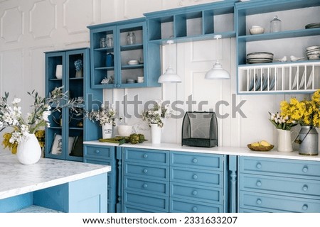 Kitchen with blue vintage furniture, wooden cupboard, kitchenware, drawers, shelves and countertop with jugs and flowers. Home decor against white walls. Monochrome interior. Royalty-Free Stock Photo #2331633207