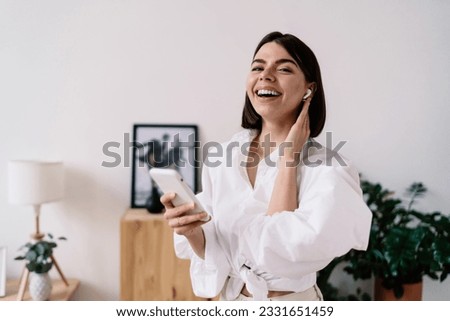 Cheerful young Hispanic woman in casual clothes laughing and talking on smartphone using wireless earphones while looking at camera at home Royalty-Free Stock Photo #2331651459
