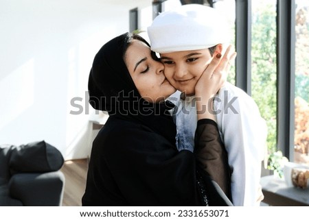 Loving Emirati parent showing love to her son at indoors. Happy Arab Parent spending time with her kid on Kandura. Middle Eastern family inside a modern house with large windows Royalty-Free Stock Photo #2331653071